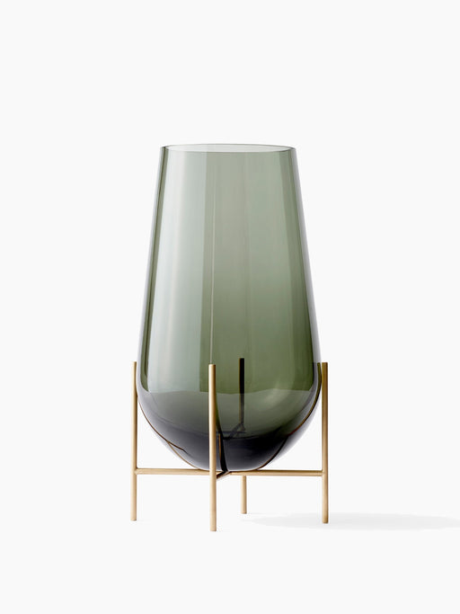 Echasse Vase M, a teardrop-shaped, smoked green glass vase on a slender, minimalist four-legged brass stand.