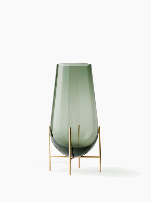 The small Echasse Vase, a smokey green glass vessel held in a slender, four-legged brass stand on a white background.