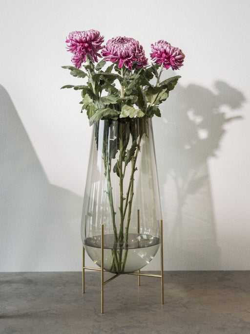 The large Echasse Vase in smoked glass on a brass stand, holding three pink chrysanthemums, on a grey surface.