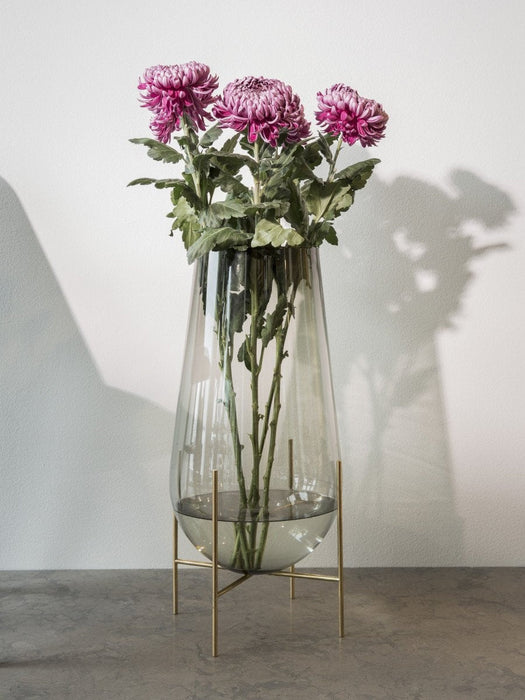 The large Echasse Vase in smoked glass on a brass stand, holding three pink chrysanthemums, on a grey surface.