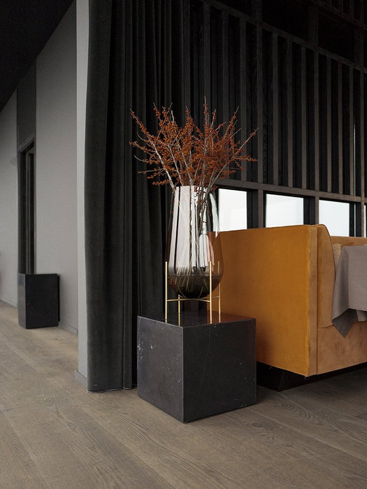 A tall, smoked glass Echasse Vase with dried branches on a brass stand, on a black marble Plinth next to a yellow sofa.