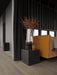 A tall, smoked glass Echasse Vase with dried branches on a brass stand, on a black marble Plinth next to a yellow sofa.