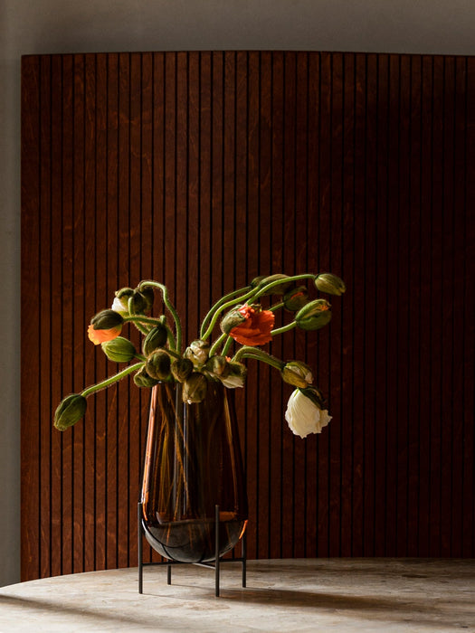 A tall, teardrop-shaped amber glass vase in a black metal stand, holding a bouquet of orange and white poppies with buds.