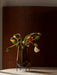A tall, teardrop-shaped amber glass vase in a black metal stand, holding a bouquet of orange and white poppies with buds.
