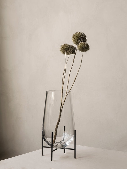 A large, clear glass Echasse Vase on a slender black metal stand, holding three dried globe thistles against a beige wall.