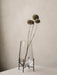 A large, clear glass Echasse Vase on a slender black metal stand, holding three dried globe thistles against a beige wall.