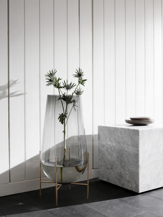 A tall Echasse Vase in smoked glass on a brass stand holds a thistle stem, next to a white marble plinth on a dark floor.