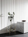 A tall Echasse Vase in smoked glass on a brass stand holds a thistle stem, next to a white marble plinth on a dark floor.