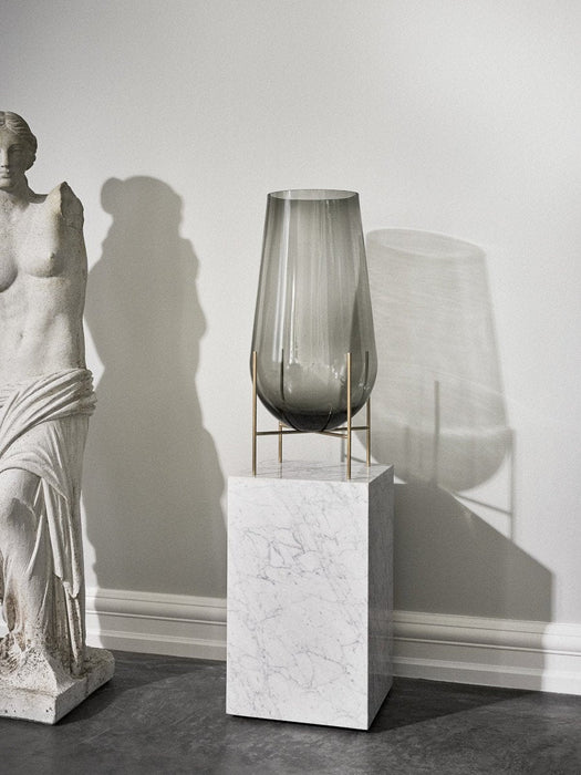 A smoked glass Echasse Vase with a brass stand sits atop a white marble Plinth in a minimalist room next to a statue.