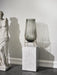 A smoked glass Echasse Vase with a brass stand sits atop a white marble Plinth in a minimalist room next to a statue.