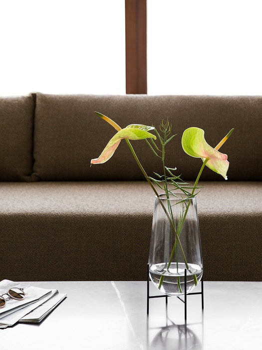 A glass Echasse Vase on a black metal stand, containing two anthurium flowers, sits on a marble table before a brown sofa.