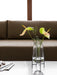 A glass Echasse Vase on a black metal stand, containing two anthurium flowers, sits on a marble table before a brown sofa.