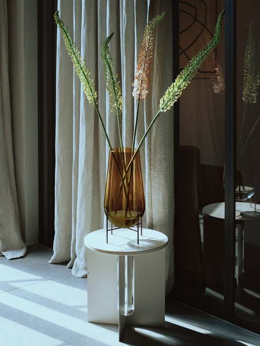 A large amber glass Hasse Vase with tall foxtail lilies sits on a white Androgyne Side Table in a sunlit room by curtains.