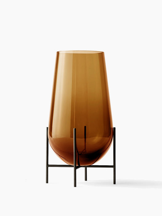 The Hasse Vase in amber glass, medium size, resting in its minimalist bronze-brass stand on a white background.