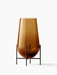 The Hasse Vase in amber glass, medium size, resting in its minimalist bronze-brass stand on a white background.