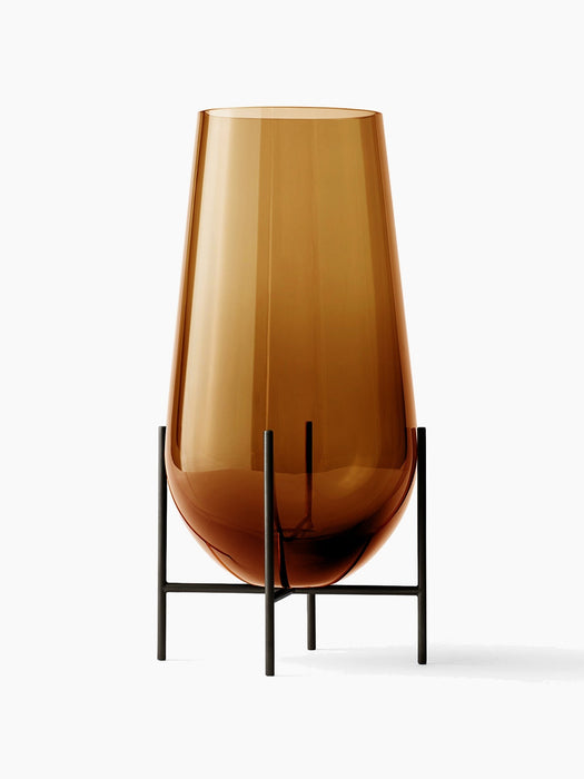A large Hasse Vase in amber-coloured glass, resting in a minimalist bronze-brass metal stand against a white background.