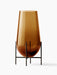 A large Hasse Vase in amber-coloured glass, resting in a minimalist bronze-brass metal stand against a white background.