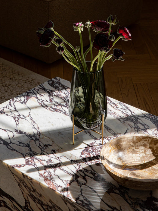 A grey glass vase with dark red ranunculus flowers on a marble coffee table, beside a wooden bowl, illuminated by sunlight.