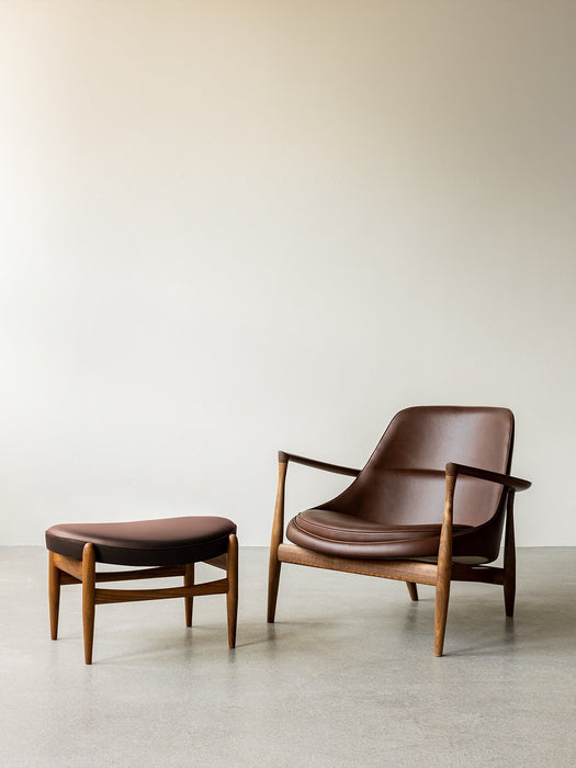 Elizabeth Lounge Chair and Ottoman in brown leather with walnut frames, set against a plain wall on a concrete floor.