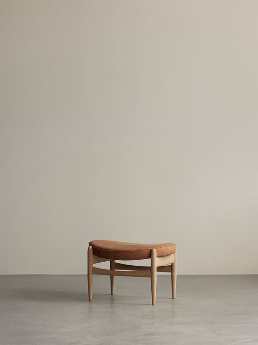 The Elizabeth Ottoman with a natural oak frame and a curved, cognac leather seat, resting on a grey concrete floor.