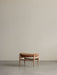 The Elizabeth Ottoman with a natural oak frame and a curved, cognac leather seat, resting on a grey concrete floor.