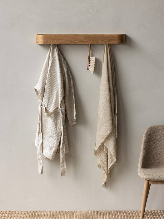 The wooden Epoch Rack mounted on a wall with a striped bathrobe, brush, and linen towel. A Harbour Chair is beside it.