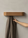 A hand hangs a brown textured throw on the wooden Epoch Rack, which is mounted on a light grey, textured wall.