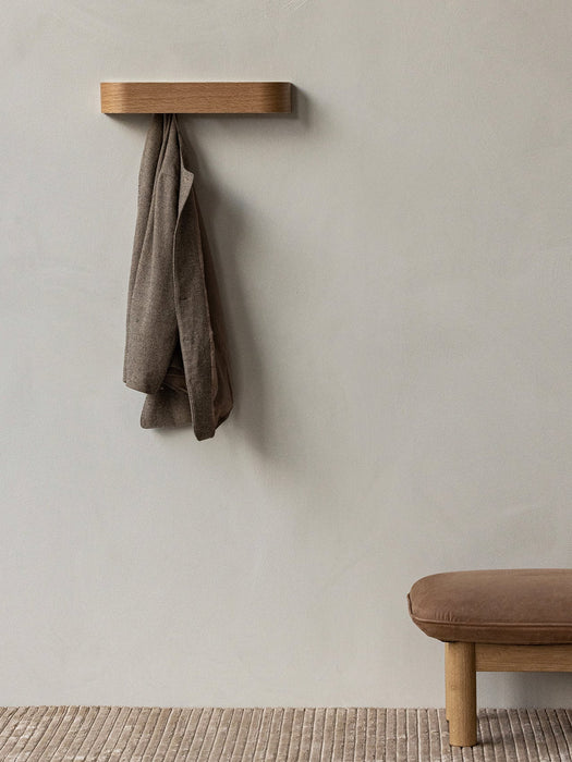 A brown jacket hangs from a wall-mounted wooden Epoch Rack. Below, a brown Brasilia Ottoman sits on a textured Houkime Rug.