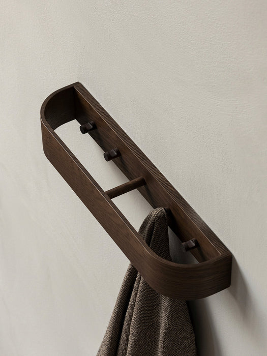 Angled view of the dark wood Epoch Rack, a modern wall rack with pegs, holding a brown woven towel against a light-grey wall.