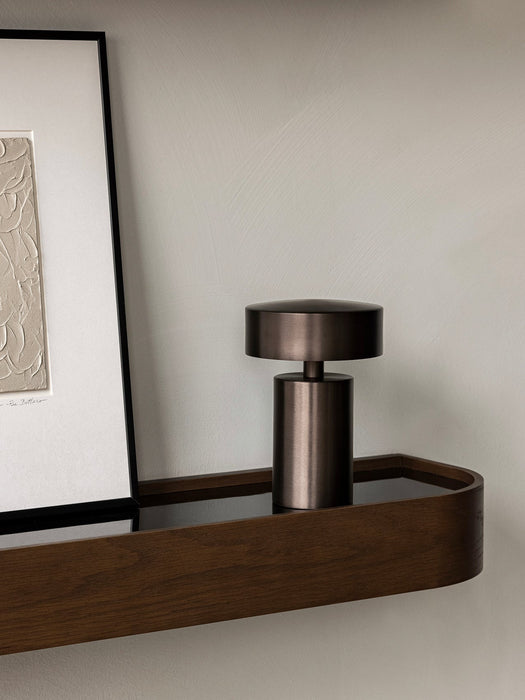 A close-up of the Column Table Lamp Portable in a dark bronze finish, placed on a dark wood Epoch Shelf next to framed art.