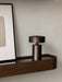 A close-up of the Column Table Lamp Portable in a dark bronze finish, placed on a dark wood Epoch Shelf next to framed art.