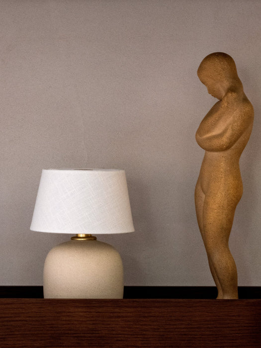 A Torso Portable Table Lamp with a textured off-white base and linen shade sits on a dark wood Epoch Shelf beside a sculpture