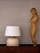 A Torso Portable Table Lamp with a textured off-white base and linen shade sits on a dark wood Epoch Shelf beside a sculpture