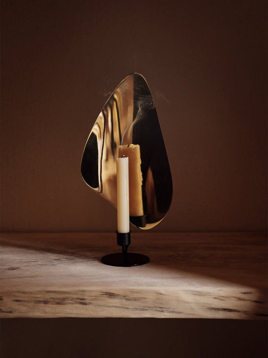 Flambeau Candle Holder sits on a rustic wooden table, its brass reflector catching the light from a single, smoking candle.