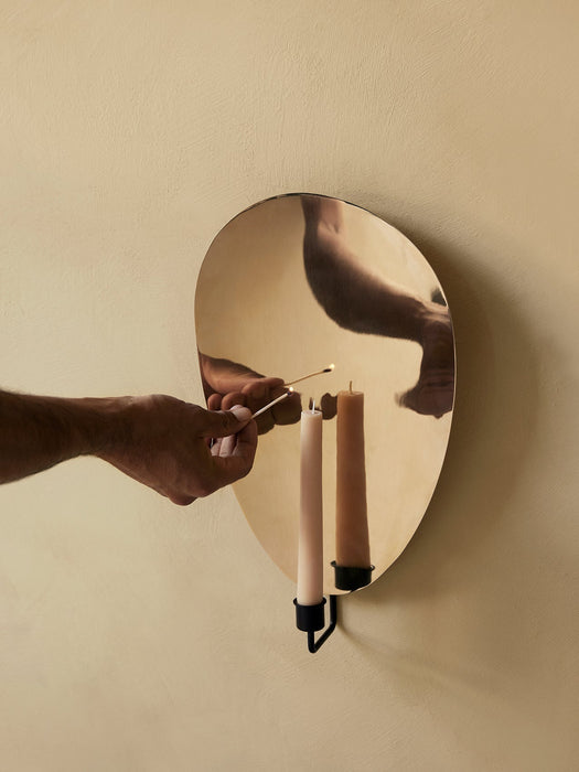 A hand lights a candle with a match in the Audo Flambeau Wall Candle Holder with a polished brass reflector on a beige wall.