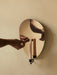 A hand lights a candle with a match in the Audo Flambeau Wall Candle Holder with a polished brass reflector on a beige wall.
