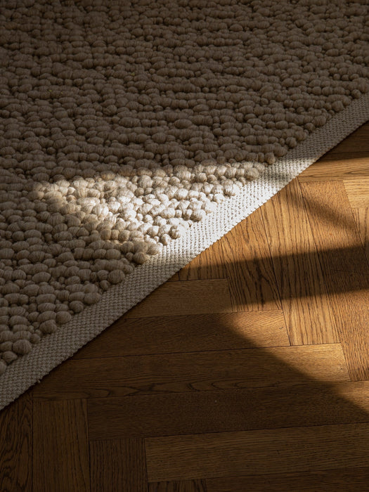 Close-up of a beige, bobbled-texture rug on a dark wooden parquet floor, highlighted by a strong ray of sunlight.