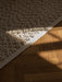 Close-up of a beige, bobbled-texture rug on a dark wooden parquet floor, highlighted by a strong ray of sunlight.