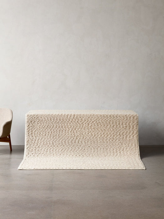 The ivory Gravel Rug draped over a plinth, showcasing its knobbly wool texture in a minimalist room with a concrete floor.