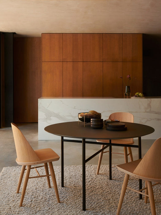 A black, round Snaregade Table and three light wood Synnes Chairs on a textured rug in a warm, modern dining space.