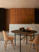A black, round Snaregade Table and three light wood Synnes Chairs on a textured rug in a warm, modern dining space.