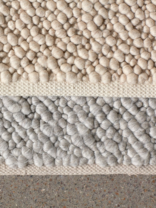A detailed close-up of the Gravel Rug, showing the soft, pebbled wool texture in ivory and light grey on a concrete floor.