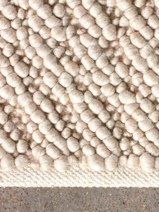 A close-up shot of the Gravel Rug in ivory, showing its bumpy, pebble-like wool texture against a grey concrete floor.