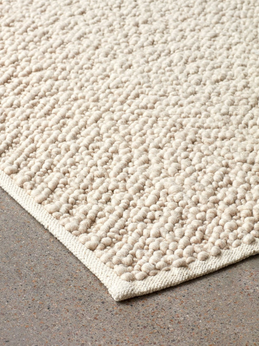 A close-up shot of the corner of the ivory Gravel Rug, highlighting its soft, bumpy wool texture on a grey concrete floor.