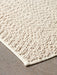 A close-up shot of the corner of the ivory Gravel Rug, highlighting its soft, bumpy wool texture on a grey concrete floor.