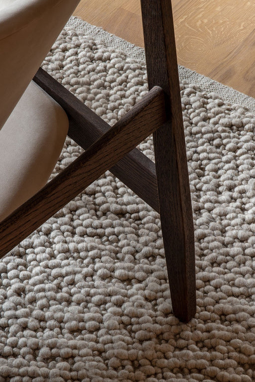 A close-up of the dark wood leg of the Knitting Chair resting on the highly textured, pebble-like weave of the Gravel Rug.
