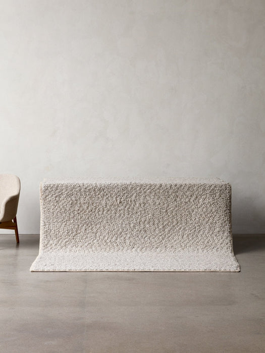 A light grey Gravel Rug with a thick, nubby texture is displayed over a platform in a minimalist room with concrete floors.