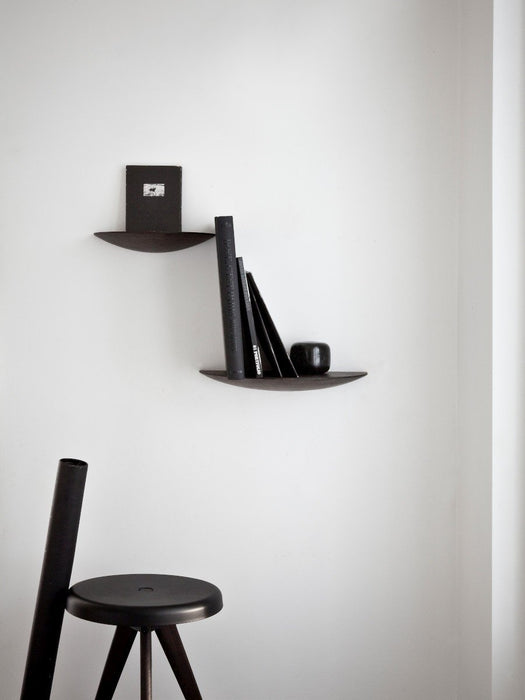 A minimalist room with two dark Gridy Fungi Shelves mounted on a white wall, styled with black books, next to a black stool.