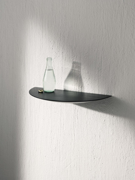 A black, leaf-shaped Gridy Fungi Shelf mounted on a white textured wall, holding a glass bottle and a small golden object.