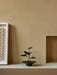 A black Hana Vase with a small anthurium plant, displayed on a minimalist shelf next to framed art against a beige wall.